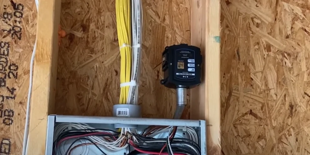 A black whole house surge protector mounted on a wooden wall next to an electrical panel with various colored wires, including yellow, white, and red, bundled together. The panel contains multiple connected wires, and the surge protector is attached with a conduit pipe.