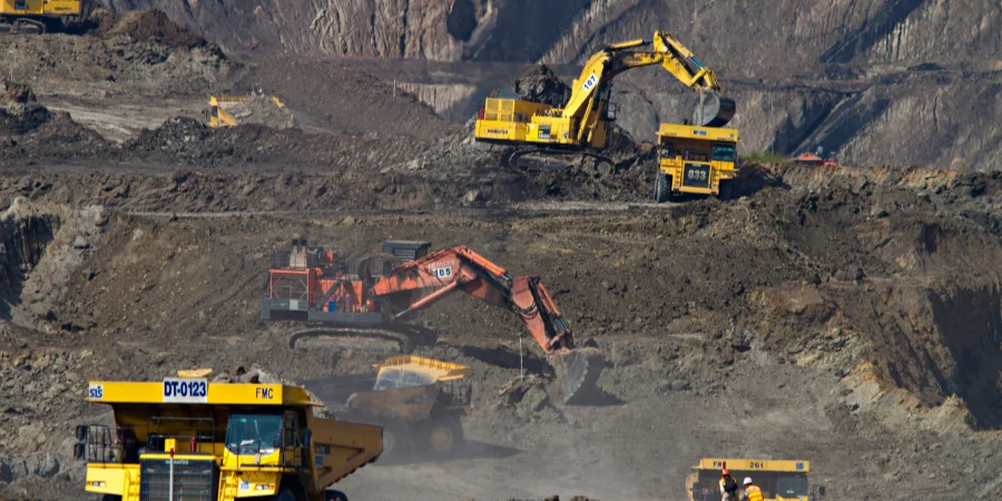 A busy open-pit mine with multiple large yellow dump trucks and excavators working amidst piles of dark earth and rocky terrain. The machinery, including a prominent truck labeled "DT-0123," is actively moving materials, with dust rising from the site. The rugged landscape features steep slopes and a variety of heavy equipment scattered across the area.