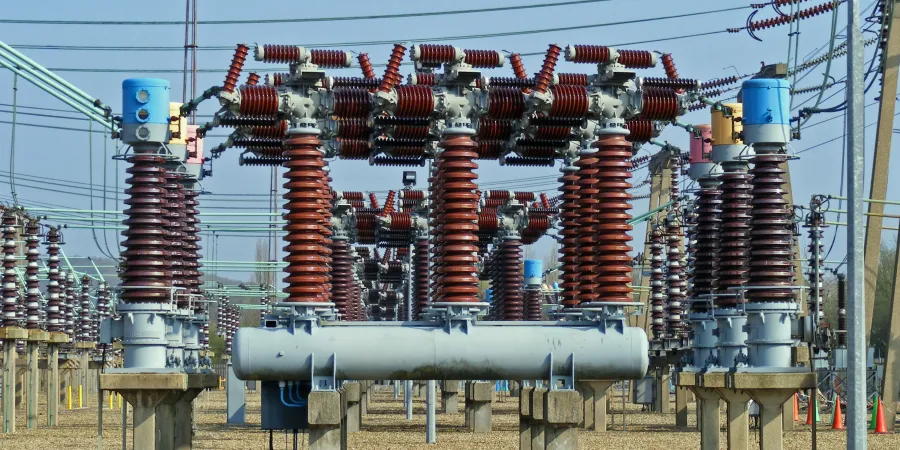An image of an air blast circuit breaker setup in an outdoor electrical substation. The scene features multiple tall, cylindrical insulators with red and gray ribbed surfaces, mounted on concrete pedestals. The breakers are connected by a network of overhead power lines and green insulators, with blue and gray control units visible at the top. In the background, additional substation equipment and power poles extend into the distance under a clear blue sky, with orange safety cones scattered on the ground.
