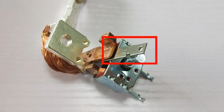A close-up view of a disassembled MCCB component on a white background, featuring a metallic assembly with a red box highlighting a bimetallic strip—a thin, curved metal piece attached to a screw. The assembly includes a bundle of copper wires and a metal bracket with multiple connection points, showcasing the thermal overload protection element within the breaker.