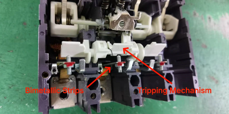 A close-up view of the internal components of a molded case circuit breaker (MCCB) on a green workbench, highlighting the bimetallic strips and tripping mechanism. Red arrows labeled