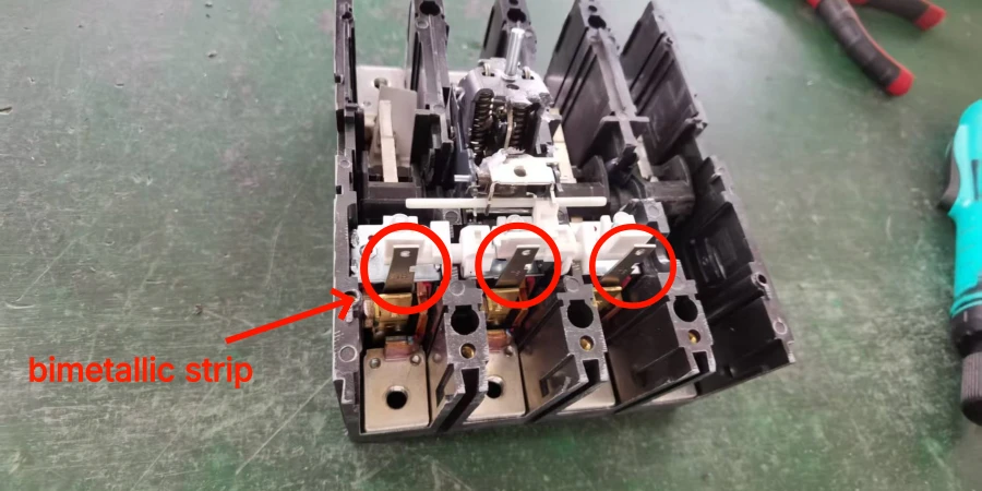 A close-up view of a partially disassembled black MCCB on a green workbench, with red circular annotations highlighting three small bimetallic strips (curved metal components) inside the mechanism. Red arrows labeled 