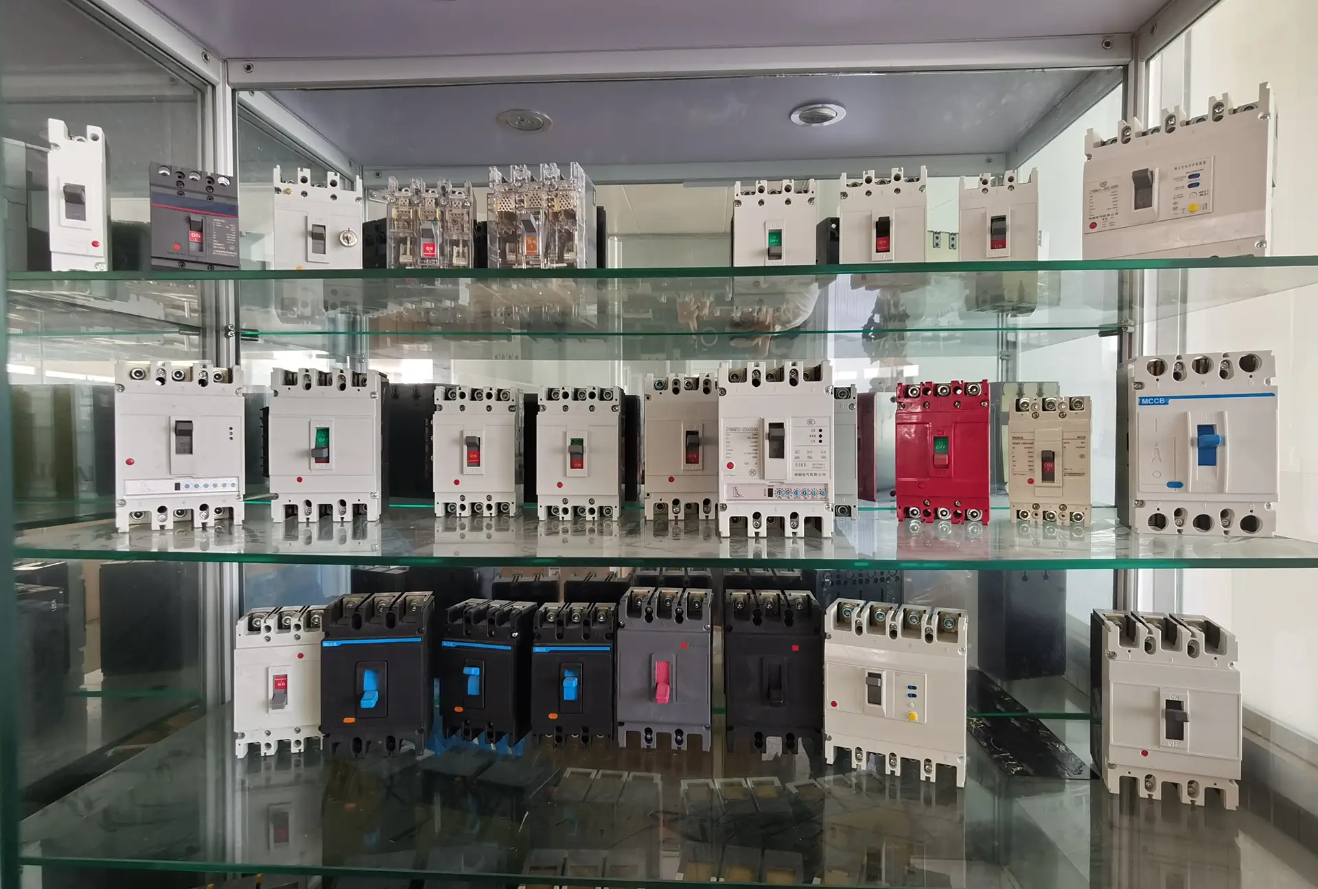 A glass cupboard showcasing a variety of Molded Case Circuit Breakers (MCCBs) arranged on multiple shelves. The breakers, in white, black, and red casings with colorful switches (green, red, blue, and pink), are neatly organized, reflecting a range of models and sizes. The background features a clean indoor setting with soft lighting, highlighting the electrical components.