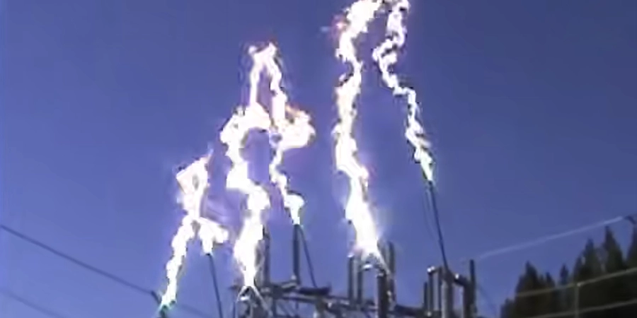 An image capturing a bright electric arc occurring on a power line structure against a clear blue sky. Multiple intense white and yellowish arcs of electricity are visible, extending from the equipment at the top of the structure, with power lines and a distant tree line in the background.