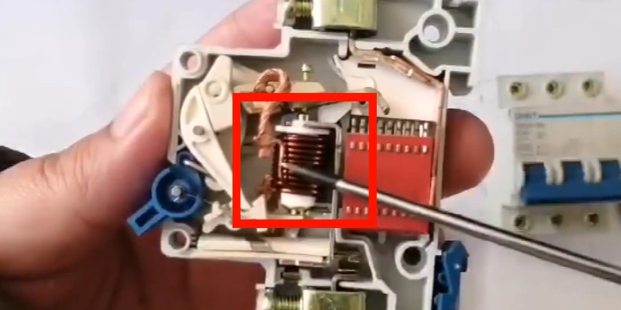 A close-up view of an opened miniature circuit breaker (MCB) held by a hand, with a red box highlighting the electromagnetic coils—a coiled copper wire assembly with a red core, mounted within the internal mechanism. A screwdriver is inserted near the coils, and the MCB's white plastic casing and blue toggle switch are visible, alongside another breaker in the background, showcasing the magnetic trip component.