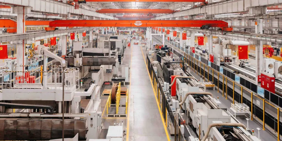 An expansive view of a modern factory floor with rows of advanced machinery and robotic arms, operating under a high ceiling with red overhead cranes. The facility is adorned with multiple Chinese flags, and the workspace is organized with yellow safety lines and railings, illuminated by bright overhead lights.