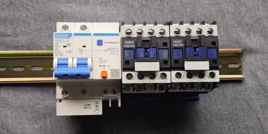 A close-up image of an electrical setup featuring a circuit breaker (MCB) and a contactor mounted on a DIN rail. The circuit breaker on the left has a blue lever and orange test button, while the contactor on the right includes multiple black and white components with blue switches. The setup is placed on a gray fabric background.