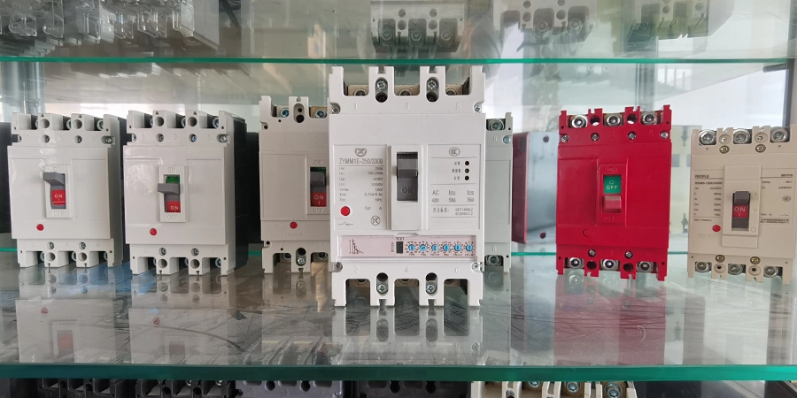 A lineup of five molded case circuit breakers (MCCBs) showcased on a glass shelf: two white 3-pole units with red toggle switches in the "ON" position on the left; a central white electronic MCCB with digital display, green indicator; a red 3-pole MCCB with green "OFF" switch; and a beige 3-pole unit with red "ON" switch on the right. Reflections visible on the glass surface below.