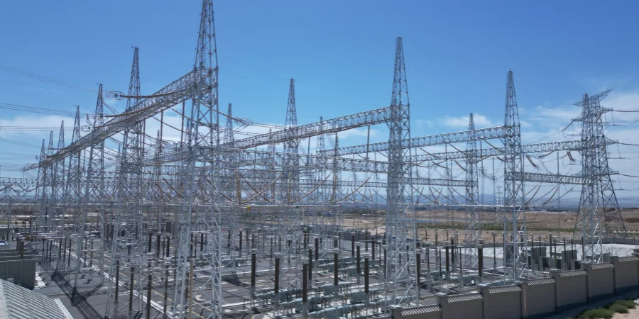 A wide view of a modern electrical substation featuring numerous tall metal lattice towers and interconnected power lines, set against a clear blue sky with some scattered clouds. The substation is surrounded by a flat, arid landscape, with a fenced perimeter and various equipment visible on the ground.