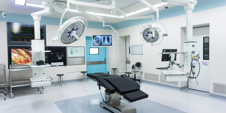 A sterile and well-equipped operating room with a central surgical table, surrounded by advanced medical monitors displaying vital signs and imaging data. Overhead, bright surgical lights are mounted on articulated arms, and various medical devices and carts are positioned around the room. The space features a clean, white design with a blue door and minimalistic furniture.