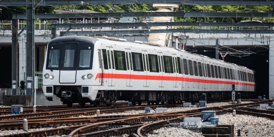 A sleek, white train with red stripes, labeled "T2860," traveling on tracks and exiting a covered station. The scene includes overhead electrical lines and a platform with structural supports, set against a backdrop of greenery and urban infrastructure.