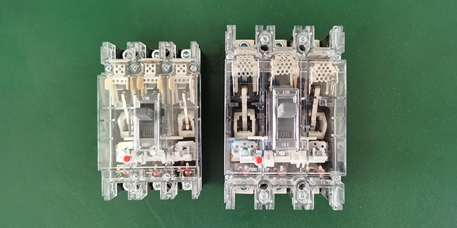 A top-down view of two molded case circuit breakers with transparent casings, placed side by side on a green background. The left breaker is smaller with an 