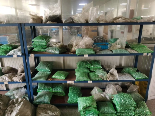 A warehouse shelf filled with green plastic bags containing screws, nuts, and similar hardware components, stacked across multiple levels. The shelves are made of blue metal, and the background shows a clean, well-lit room with additional equipment and storage visible through a glass partition.