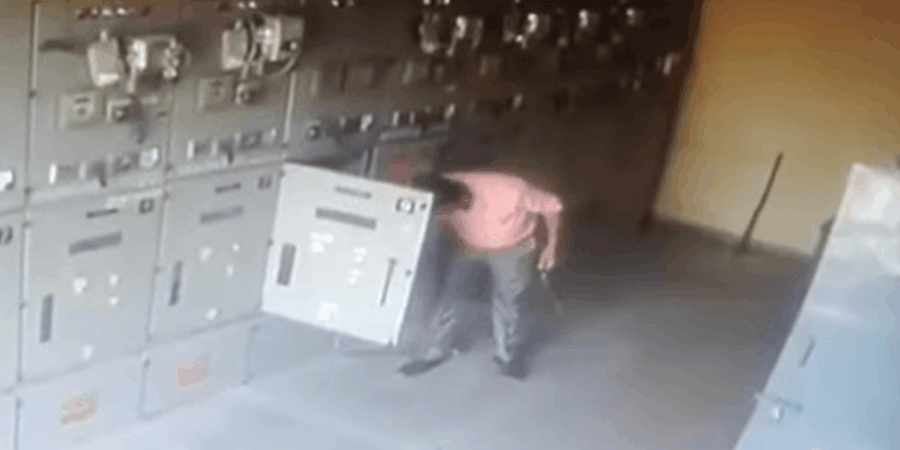 A video frame from a dimly lit electrical control panel showing a sudden arc flash explosion, with bright orange flames and sparks bursting from the center while someone appears to be inspecting or working nearby, highlighting the dangers of electrical maintenance.