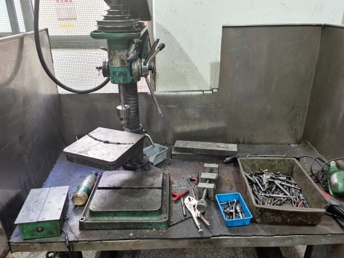 A close-up view of an old green bench drill press mounted on a sturdy metal workbench in an industrial workshop. The drill features a tall column with a hand lever and chuck, positioned over a large work table cluttered with metal scraps, a small tray of screws and nails, wrenches, pliers, and a green oil can. The background shows stainless steel panels and a window with safety mesh.