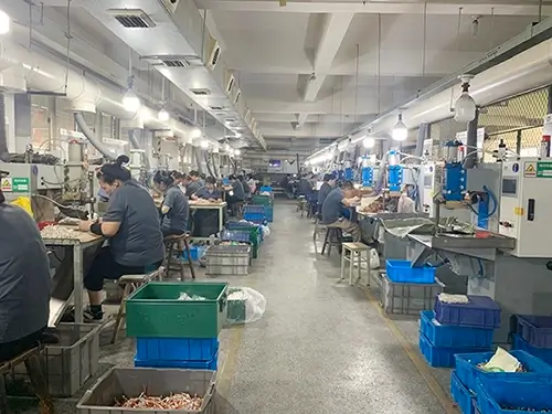 A spacious spot welding workshop with multiple workers at workstations, operating spot welding equipment and handling metal components. The room is well-lit with overhead lights, featuring blue and green storage bins, worktables, and various equipment along the sides.
