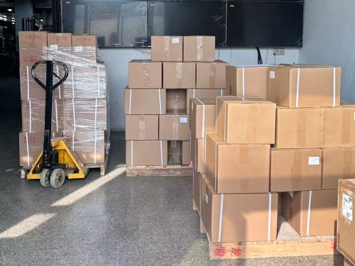 A warehouse scene featuring multiple stacks of brown cardboard boxes, some wrapped in plastic, with a yellow pallet truck parked nearby. The boxes are arranged on wooden pallets, and a large screen or window is visible in the background on a tiled floor.