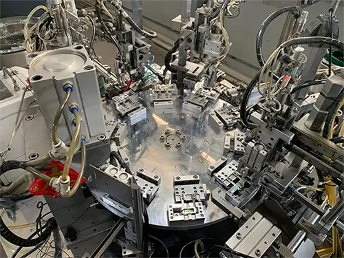 A close-up view of an automated riveting machine with a central rotating platform, surrounded by multiple robotic arms, sensors, and metallic components. The machine features various wires and hoses, operating in a clean industrial environment.