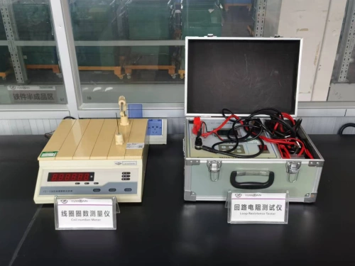 A side-by-side display of two electrical testing instruments on a black lab bench, illuminated by soft overhead light in an industrial workshop with glass partitions and shelving in the background. On the left is a compact beige coil turn counter with a digital LCD readout showing measurement values, a detachable blue probe arm on a swivel base, and a power adapter plugged into the side; labeled "Coil Turn Counter" on a white placard in Chinese and English. On the right is a rugged silver aluminum carry case, open to reveal a portable contact resistance tester with coiled red and black test leads, battery clips, a digital multimeter-style interface, and foam-padded compartments for accessories. These tools are essential for verifying electrical integrity in circuit breaker components like coils and contacts, ensuring safety and reliability in manufacturing quality assurance.