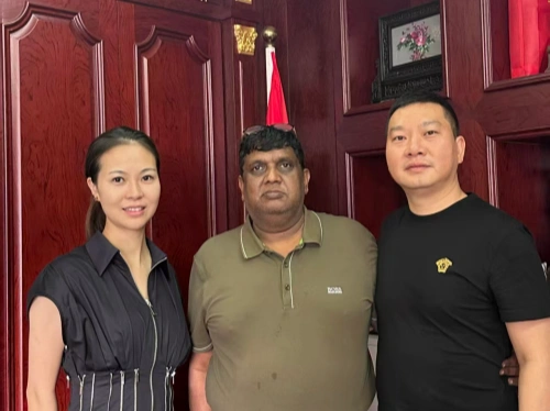 Three individuals standing together in a warmly lit room with dark wood-paneled walls, and MCB. A woman on the left wears a black dress with a unique sleeve design, a man in the middle wears a green polo shirt, and a man on the right wears a black Versace shirt. A small flag and a framed picture with flowers are visible in the background.