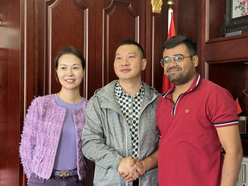 A photo of three individuals standing in front of a wooden paneled wall with a flag in the background. The person on the left is a woman wearing a purple top with a fringed jacket and a belt. The person in the middle is a man in a gray jacket over a checkered shirt, shaking hands with the person on the right. The person on the right is a man wearing a red polo shirt with white trim, also shaking hands. The setting appears to be an office with formal decor.