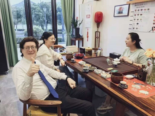 A photo of three individuals seated around a wooden table in a room with green curtains and a scenic outdoor view through the window. The person on the left is a man with glasses, wearing a white shirt and blue tie, giving a thumbs-up gesture. In the middle is a woman in a white shirt, smiling towards the camera. On the right is another woman in a light-colored top, looking sideways. The table is set with various tea cups, teapots, snacks, and a red decorative mat, with a red lantern and plants in the background, suggesting a traditional Chinese tea setting.