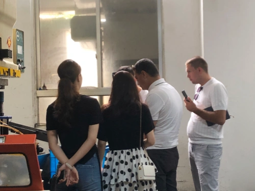A photo taken from behind of four individuals standing in a bright industrial room, observing through a large window or directly watching machinery in operation. From left to right: a woman with long dark hair tied in a ponytail wearing a black top and jeans, hands clasped behind her back; another woman with long dark hair in a black top and a black-and-white polka dot skirt, carrying a small white crossbody bag; a man in a white shirt facing slightly left; and another man in a white shirt holding a phone in one hand and a clipboard in the other. On the left side of the image, a mechanical press machine with yellow caution labels is visible, along with a blue plastic factory bin used for holding finished parts. Sunlight streams through the window, illuminating the clean industrial workspace.