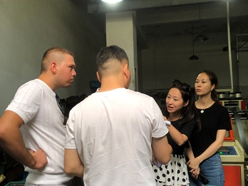 A photo of four individuals engaged in conversation in an industrial setting with machinery and equipment in the background. From left to right: a man with a buzz cut wearing a white t-shirt, standing with hands on hips and facing right; another man viewed from behind in a white t-shirt; a woman with dark hair tied back, wearing sunglasses on her head, a black top, and a black-and-white polka dot skirt, gesturing with her hand; and a woman with long dark hair in a black top and jeans, holding a phone. The room has white walls, overhead lights, and various machines and tools visible.