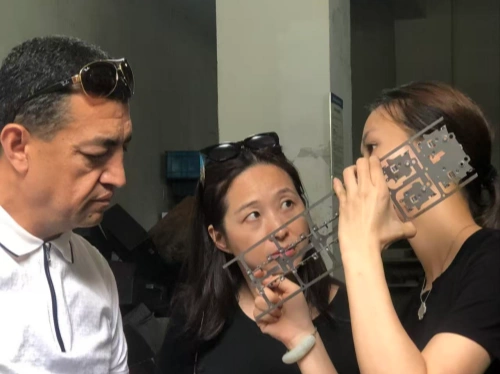 A photo of three individuals in an indoor workshop setting engaged in a product discussion. On the left, a male customer with short dark hair and sunglasses pushed up on his forehead, wearing a white collared shirt, listens attentively. In the middle, a female factory staff member with shoulder-length dark hair and sunglasses on her head, dressed in a black top, is explaining the product’s manufacturing process. On the right, another female staff member with dark hair tied in a ponytail, also in a black top, is holding a perforated metal component, using it to illustrate a detail of the process. Blurred workshop equipment and posters are visible in the background, emphasizing an active industrial environment.