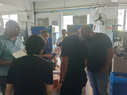 A photo taken in a factory environment, with machinery, pipes, and blue cabinets visible in the background. On the left, a man in a light blue polo shirt holds a notebook and appears to be explaining. Next to him, a person viewed from behind with short dark hair in a black top is pointing forward. In the middle, a man in a light-colored shirt looks down, while several others in blue uniforms stand in the back. On the right, two men in dark t-shirts are leaning over to inspect copper wires on the floor, carefully observing production details. The environment includes fans, control panels, and a blue bin, illustrating a tour of the factory production process.