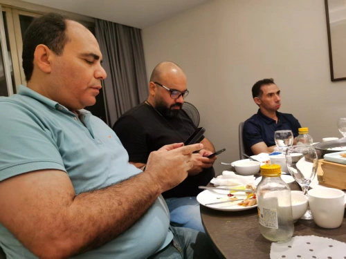 A photo of three men sitting at a round wooden table in an indoor setting with beige walls and curtains. The man on the left, bald with a beard, wearing a light blue polo shirt, is intently looking at his phone held in both hands. The middle man, bald with glasses, in a black t-shirt, is also focused on his phone. The man on the right, with short dark hair, in a navy blue shirt, holds a glass while looking forward. The table is cluttered with plates of food remnants, bowls, utensils, a bottle of yellow sauce, a water bottle, glasses, and cups, indicating a casual meal in progress.