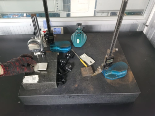 A close-up view of a granite surface plate in an industrial metrology lab, with a hand wearing a red knitted glove and green wristband holding a small labeled metal sample with QR code. Nearby are a blue Mitutoyo digital depth gauge, chrome height gauges on adjustable stands, scattered black clamps, brass blocks, a yellow-handled screwdriver, and blue plastic components on the dark surface. The setup is enclosed in a glass-walled room with overhead lighting.