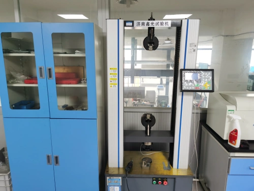 An industrial laboratory featuring a tall, white-framed electronic universal testing machine at the center, equipped with a black hydraulic gripper arm suspended from the top crosshead, a digital control monitor displaying interface options, and yellow safety warning labels on the transparent safety enclosure. To the left, two tall blue metal storage cabinets with glass doors hold organized lab supplies including red toolboxes, plastic bins, documents, and small equipment. The right side shows a gray workbench with additional tools like a drill, clamps, and a red-labeled cleaning spray bottle next to a white lab cart. The cleanroom environment includes overhead fluorescent lighting, wire mesh guards, and background machinery visible through large windows。