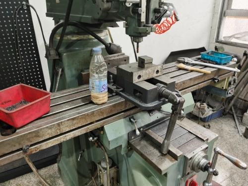 A green industrial milling machine with a water bottle and a red tray of metal shavings on its table, featuring a drill bit and coiled air hose, set in a cluttered workshop with tools and equipment in the background.
