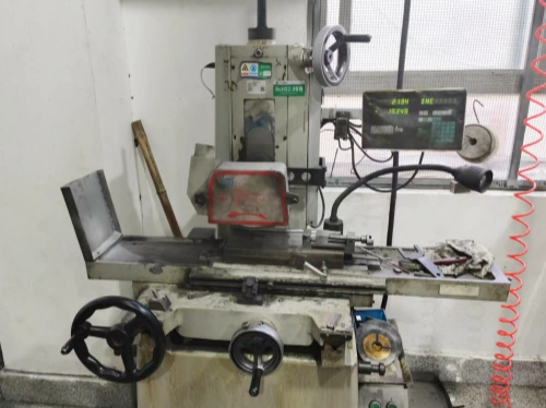 An industrial surface grinder machine in a workshop, featuring a sturdy metal frame with a large handwheel, a digital display, and a grinding wheel enclosed in a red safety guard. The machine is equipped with a worktable, control knobs, and a coiled red power cord. A wooden handle and various metal tools are placed nearby, with a window and safety mesh in the background.