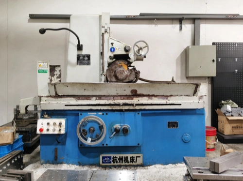 A blue industrial surface grinding machine with visible wear and metal shavings scattered around, situated in a workshop with a white wall and various tools in the background.