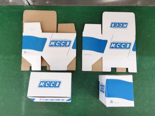 A green workbench displaying four cardboard boxes designed as inner color packaging, featuring a blue and white color scheme with "MCCB" branding. The boxes include two partially folded rectangular boxes and two fully formed boxes, one rectangular and one cubic.