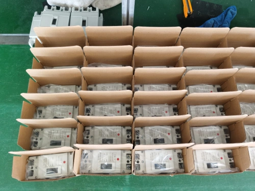 A green assembly line workstation displaying multiple open white inner packaging boxes, each containing a white mccb unit sealed in a plastic bag. The inner boxes are arranged neatly in rows, with additional electrical components and tools visible in the background.