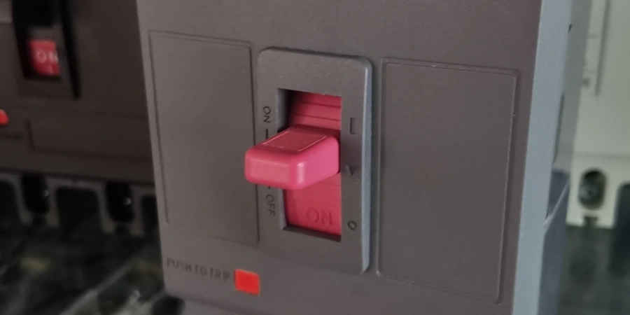 Close-up of a gray MCCB with pink handle in the middle tripped position between ON and OFF. Labels show ON at top, OFF at bottom, and red 'PUSH TO TRIP' button below.
