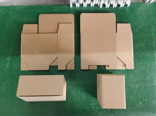 A green assembly line surface displaying several unfinished neutral inner packaging boxes made of Kraft paper, including two partially folded rectangular boxes, one fully assembled rectangular box, and one cubic box. Workshop equipment and tools are visible in the background, showing the packaging preparation stage before final product assembly.