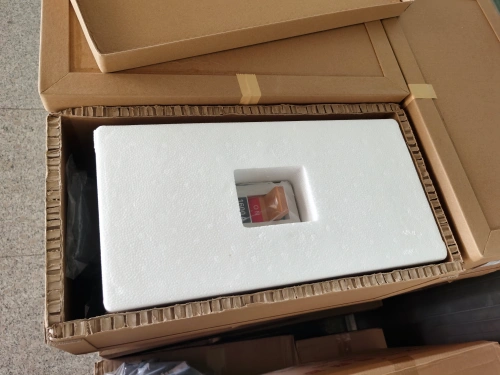 An open brown cardboard box containing a white foam padding insert, internally securing a high-current MCCB, featuring a white housing. Additional stacked boxes are visible in the background, placed on the floor.