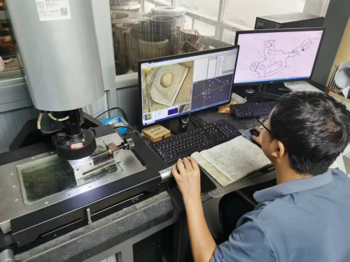 A detailed view of a modern industrial metrology lab where an Asian male technician in a light blue collared shirt and glasses sits focused at a sleek black workstation, operating an optical coordinate measuring machine (CMM). The tall, silver cylindrical CMM stands prominently on the left, featuring a precision stage with a glass worktable illuminated from below, a red-knobbed objective lens turret, and adjustable arm for non-contact scanning. On the dual-monitor setup to the right, the left screen displays a high-magnification live image of a circular metal workpiece with overlaid digital grids, measurement points, and color-coded analysis tools, while the right screen shows a detailed CAD blueprint of mechanical components including gears and brackets. Surrounding the setup are a mechanical keyboard, a worn notebook filled with hand-drawn engineering sketches, a small wooden storage box, and scattered tools on a granite surface base for vibration damping. In the background, large windows reveal a workshop with wooden barrels, shelving units, and diffused natural light, highlighting a collaborative environment for quality control and dimensional inspection in manufacturing.
