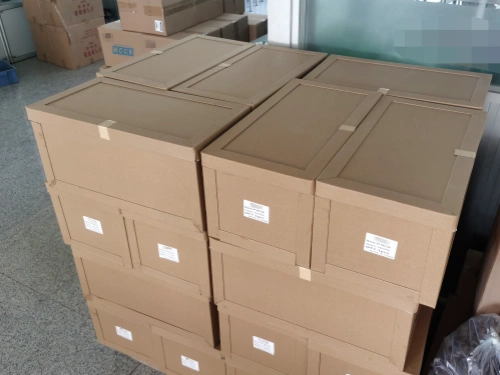 A stack of brown cardboard outer boxes with corner protectors, neatly arranged in a warehouse setting. The boxes are labeled and taped, with additional boxes and packaging materials visible in the background on a tiled floor.