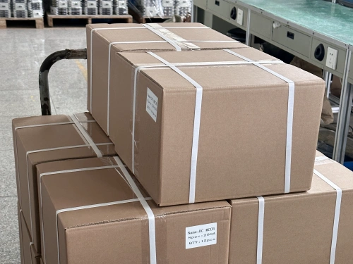 A stack of brown cardboard outer cartoons secured with white strapping, placed on a cart in an industrial warehouse. The boxes are labeled with text, and additional boxes and equipment are visible in the background on a tiled floor.