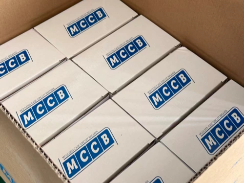 An open brown cardboard outer box containing multiple rows of white inner packaging boxes, each prominently labeled "MCCB" in blue text, arranged neatly inside.