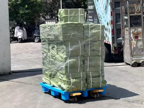 A tall stack of goods wrapped in green woven sacks, secured on a blue wheeled pallet in an outdoor loading=