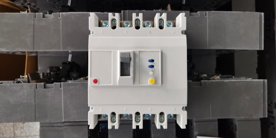 White RCCB sitting on top of stacked gray MCCB bases. Gray handle in ON position, red light on left, yellow light below, two blue lights on right. Terminals labeled 1,3,5,N on top and 2,4,6,N on bottom.