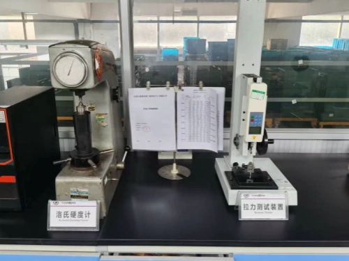 A laboratory workbench setup featuring two industrial testing instruments side by side against a blurred background of shelving and storage in a manufacturing facility. On the left is a gray Rockwell hardness tester, with a large analog dial gauge displaying measurements, a vertical drill-like indenter arm, and a sturdy base clamp; it's labeled "Rockwell Hardness Tester" on a white placard. To the right is a modern white digital tensile testing machine, equipped with a vertical column, electronic control panel with LCD display, and a clamp mechanism for pulling samples; labeled "Tension Tester" on a similar placard. Between them sits an open binder of printed test data sheets with tables of measurements and Chinese annotations, clipped to a stand, alongside a small metal disc on a pedestal. The black countertop and glass enclosure emphasize precision engineering for material strength evaluation in electrical component production.