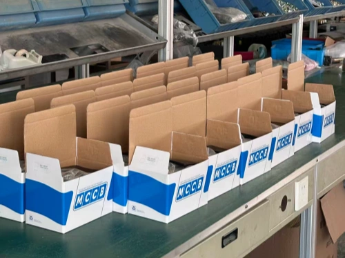 A photo showing a row of open cardboard boxes labeled "MCCB" with blue and white branding, neatly arranged on an assembly workstation in an industrial setting. Some boxes contain items inside. In the background, shelving units with blue bins and various materials are visible, indicating a workshop or production environment.