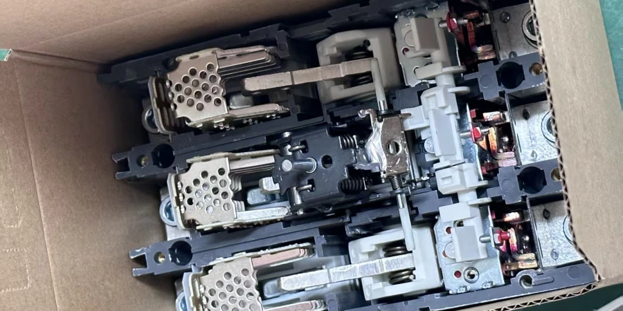 A cardboard box containing three semi-assembled MCCB mechanisms, featuring black and gray plastic frames with metallic components, including perforated plates, springs, and red-tipped connectors, designed for molded case circuit breaker functionality.